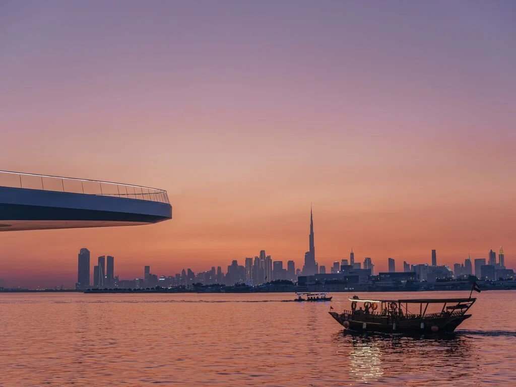 A city skyline at sunset with tall skyscrapers, including the Burj Khalifa, viewed across a body of water with a boat in the foreground.