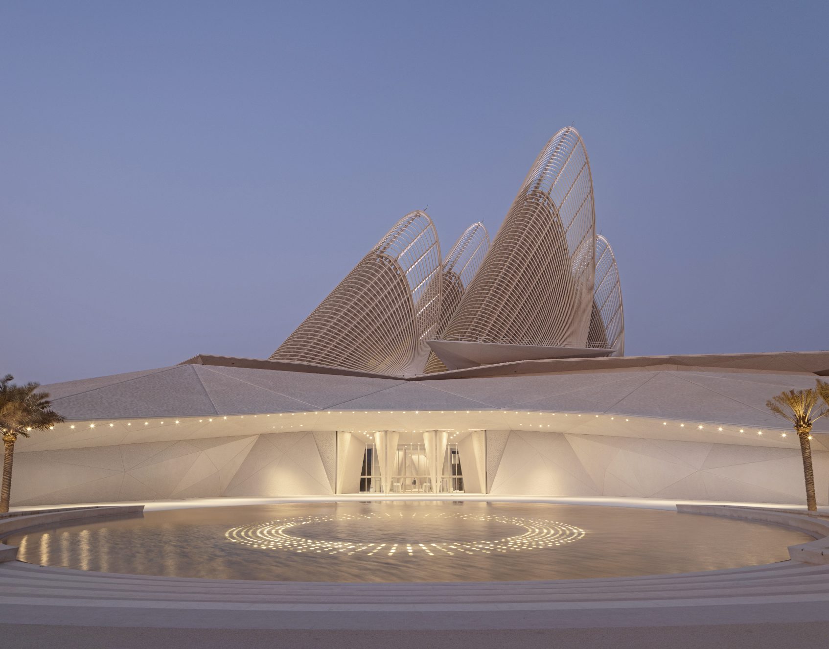 Modern architectural building with curved, ribbed design against a clear blue evening sky, illuminated at the base with string lights and surrounded by a circular water feature and palm trees.