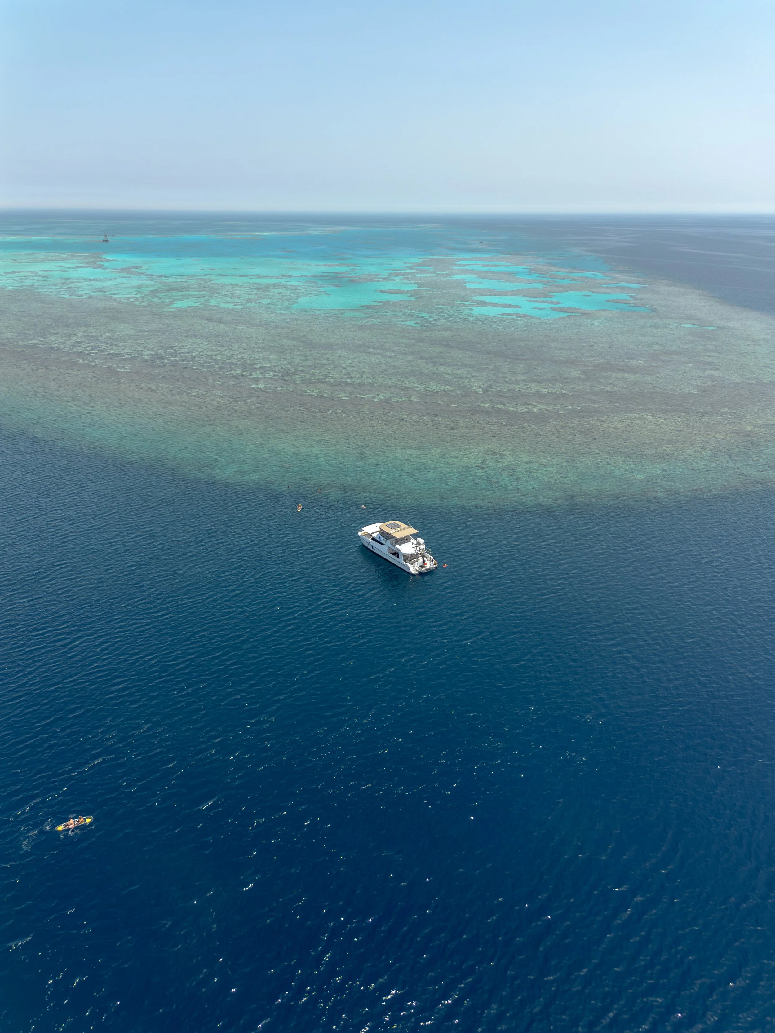 Aerial view of the ocean showing a yacht, a small boat with two people, and a paddleboard with two people, with turquoise water and patches of coral reef visible.