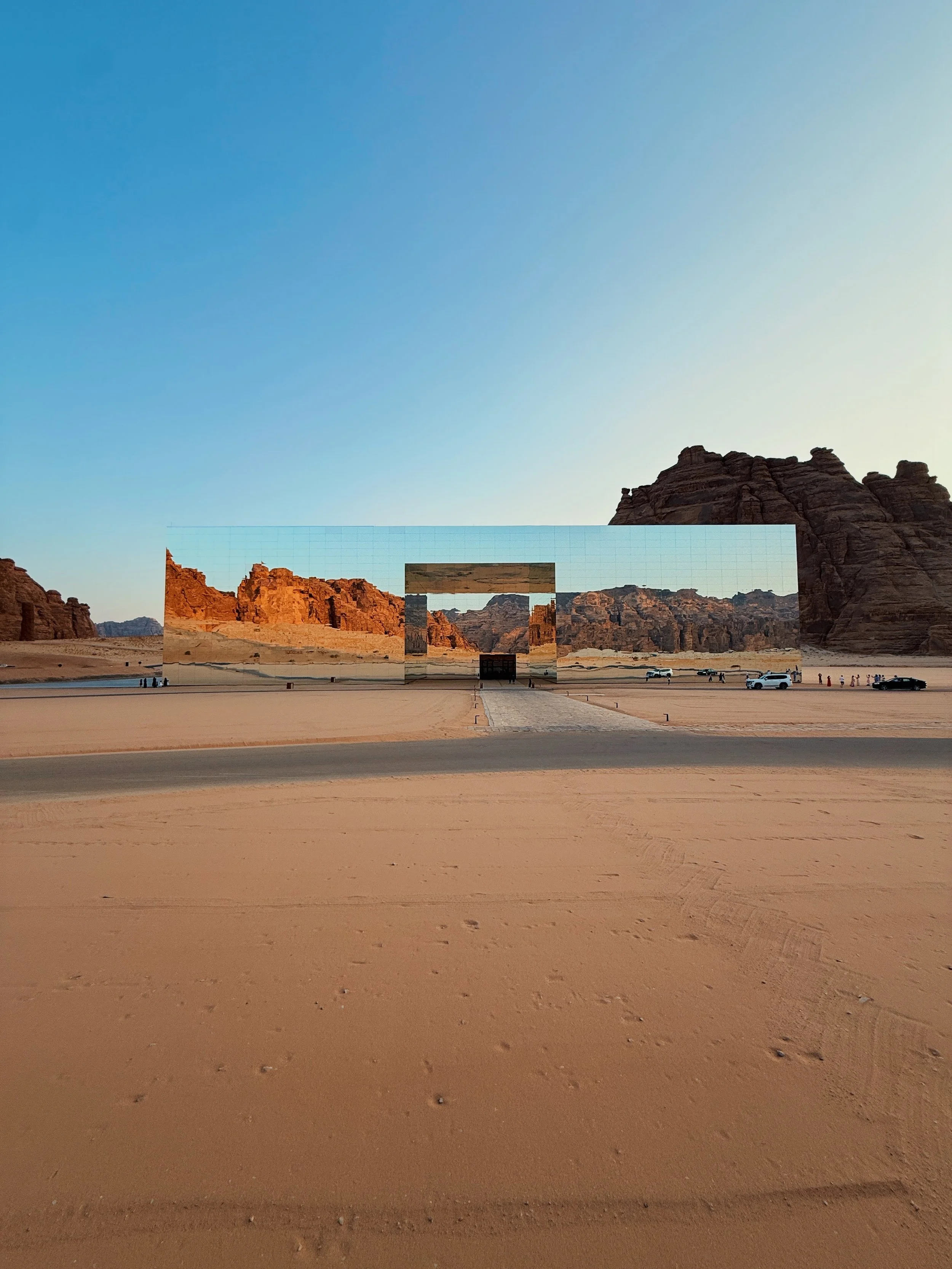 A desert landscape with rocky formations in the background, a large reflective mirror installation in the middle, and a few cars and people in the distance.
