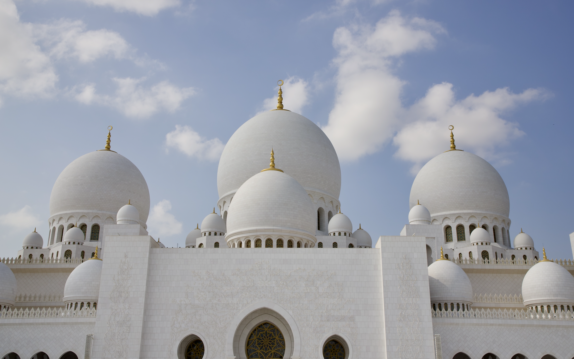 White marble mosque with multiple domes and gold finials against a partly cloudy sky.