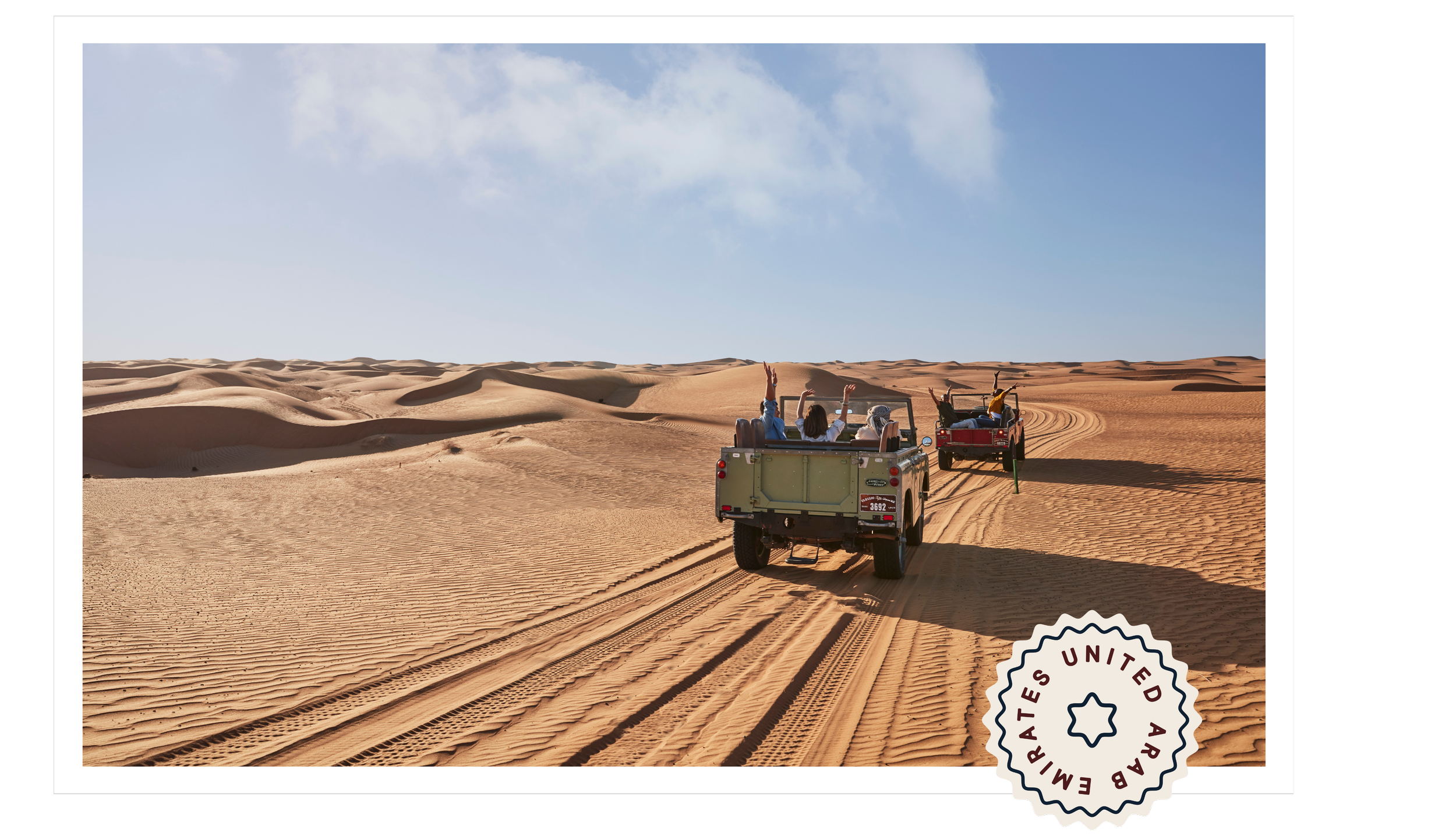 Two open-air jeeps driving through a vast desert with sand dunes under a blue sky, with three people in each jeep enjoying the ride.