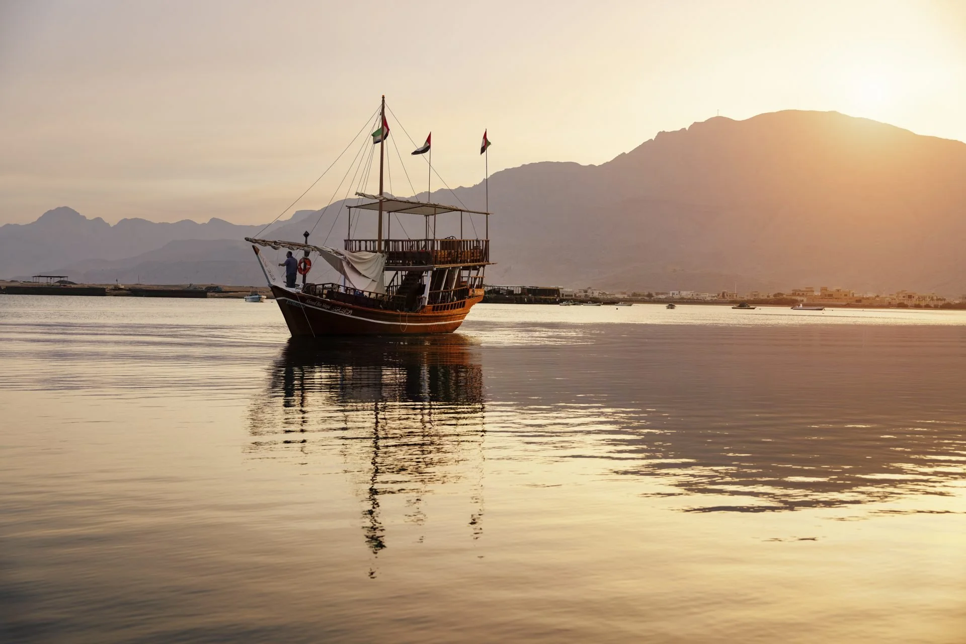 A wooden boat floating on calm water during sunset or sunrise with mountains in the background and flags flying from the boat's mast.