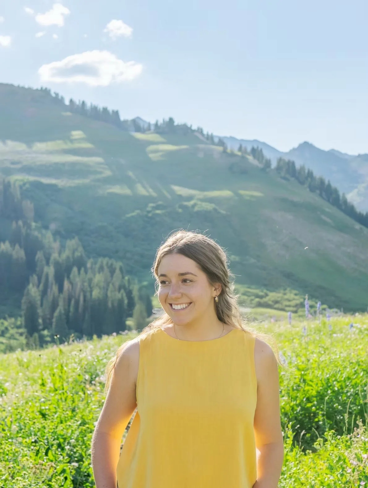 A woman in a yellow sleeveless top standing in a green field with mountains in the background under a sunny sky.