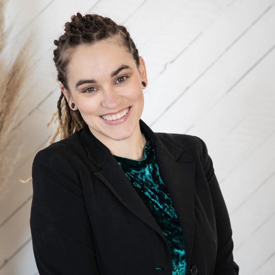 A young woman with light skin and dreadlocks smiling at the camera, dressed in a black blazer with a green velvet top, standing in front of a white wooden plank wall with some pampas grass in the background.