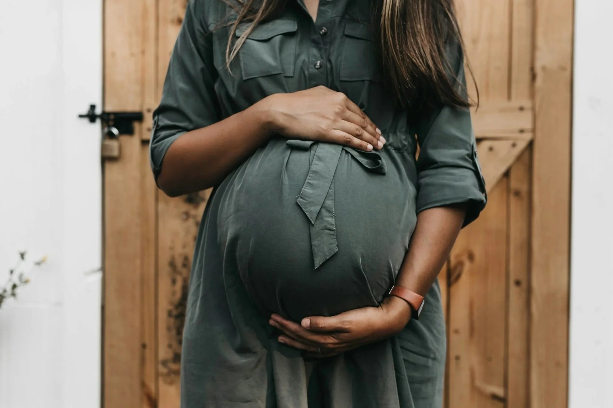A pregnant woman in a green dress gently cradles her belly with both hands, standing in front of a wooden background.