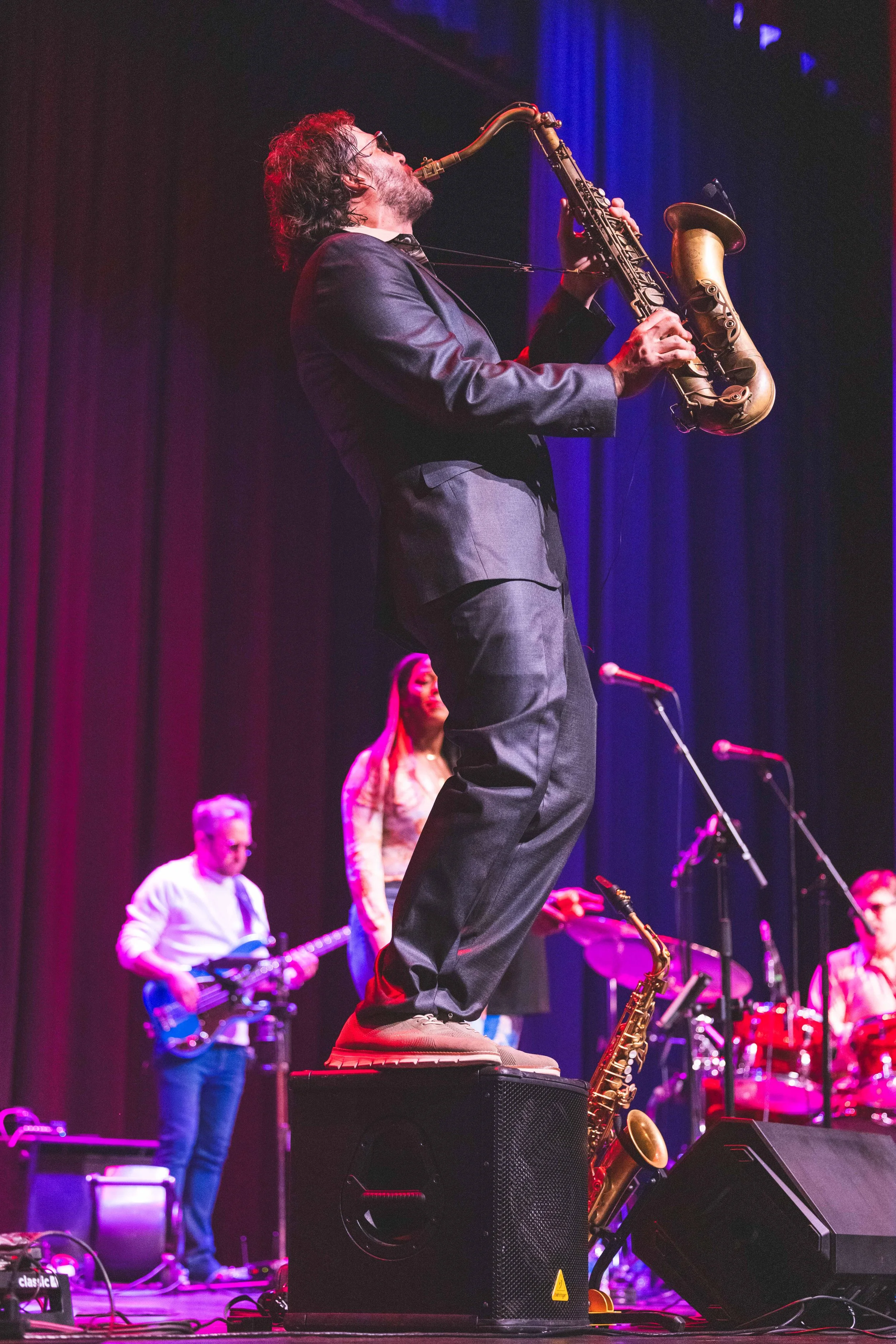 Musician standing on a speaker playing a saxophone during a live concert, with other band members in the background.