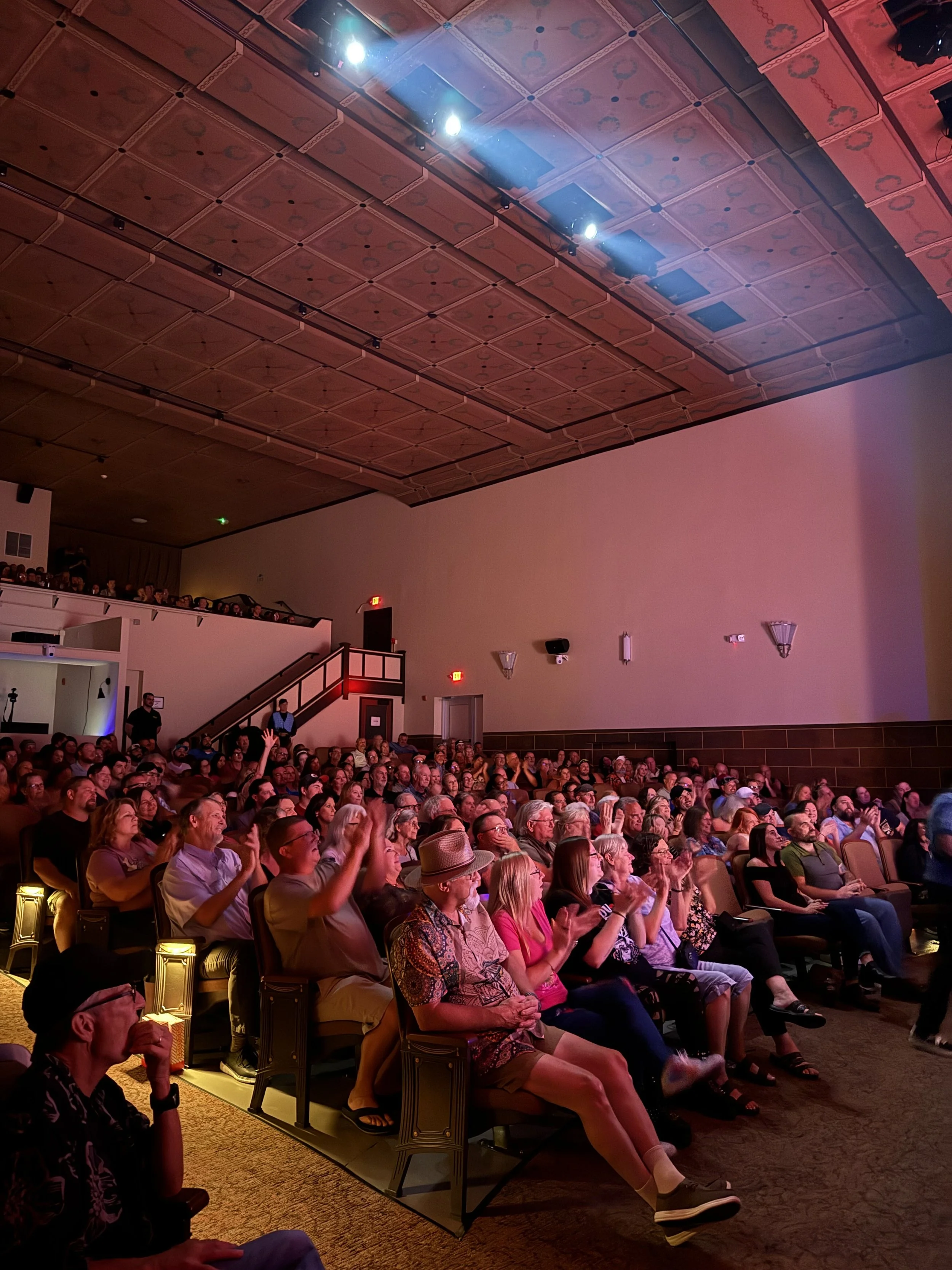 Audience seated indoors, watching a performance on stage in a theater with a high ceiling and wood paneling.