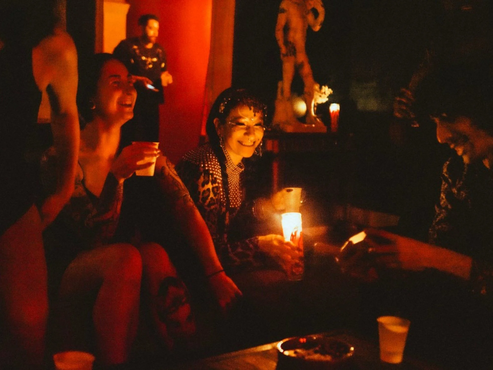 People sitting around a table in a dimly lit room, celebrating with drinks and candles, with some standing and others smiling.