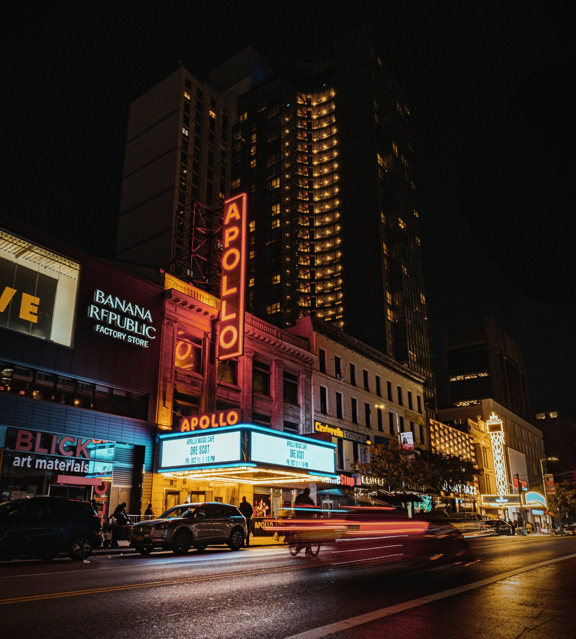Nighttime city scene with illuminated storefronts and a high-rise building. Neon signs for Apollo and other businesses are visible. Cars and pedestrians are on the street, with light trails from moving vehicles.