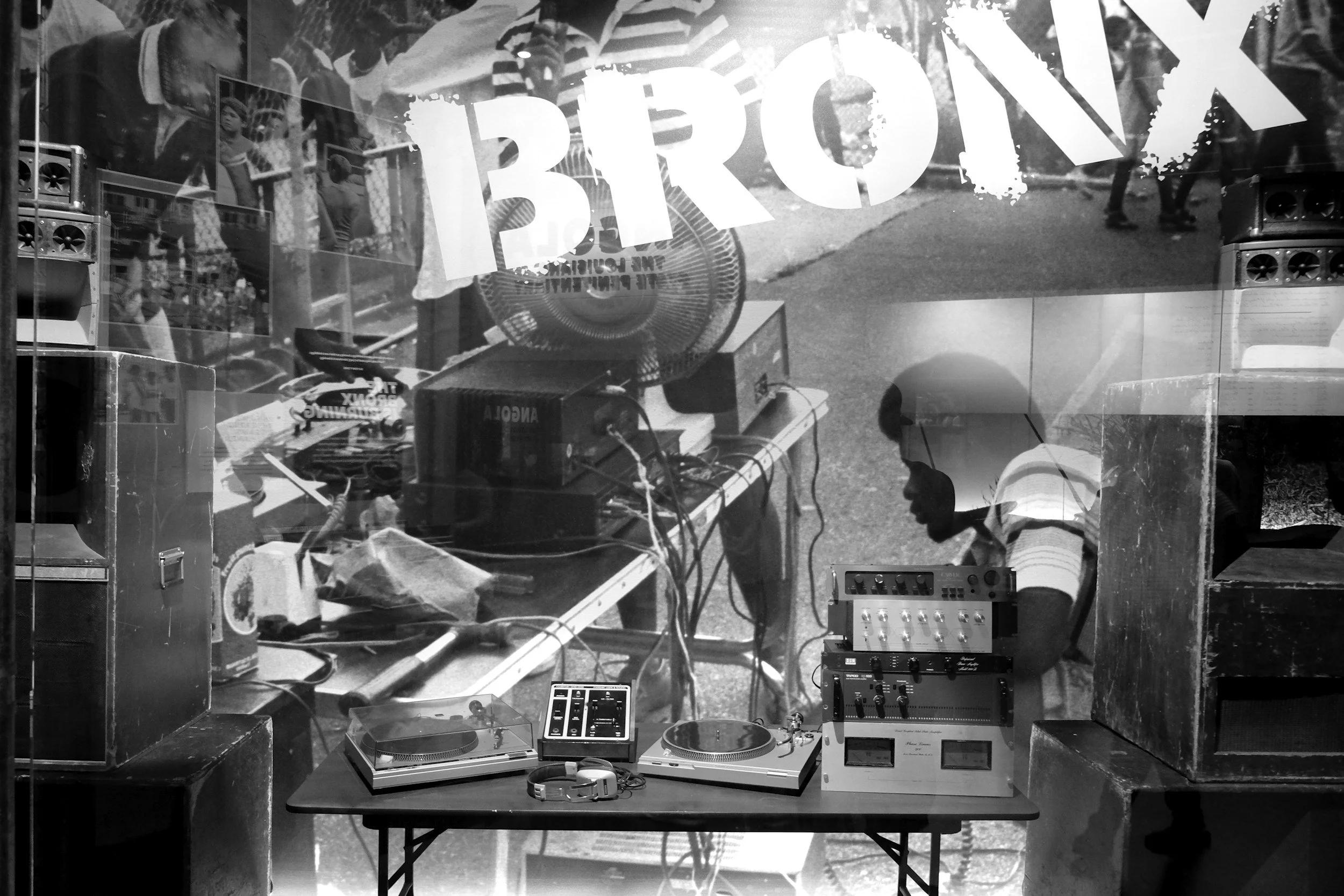 A view through a glass window showing a table with DJ equipment including turntables, a mixer, and headphones, as well as posters and people in the background on the street.
