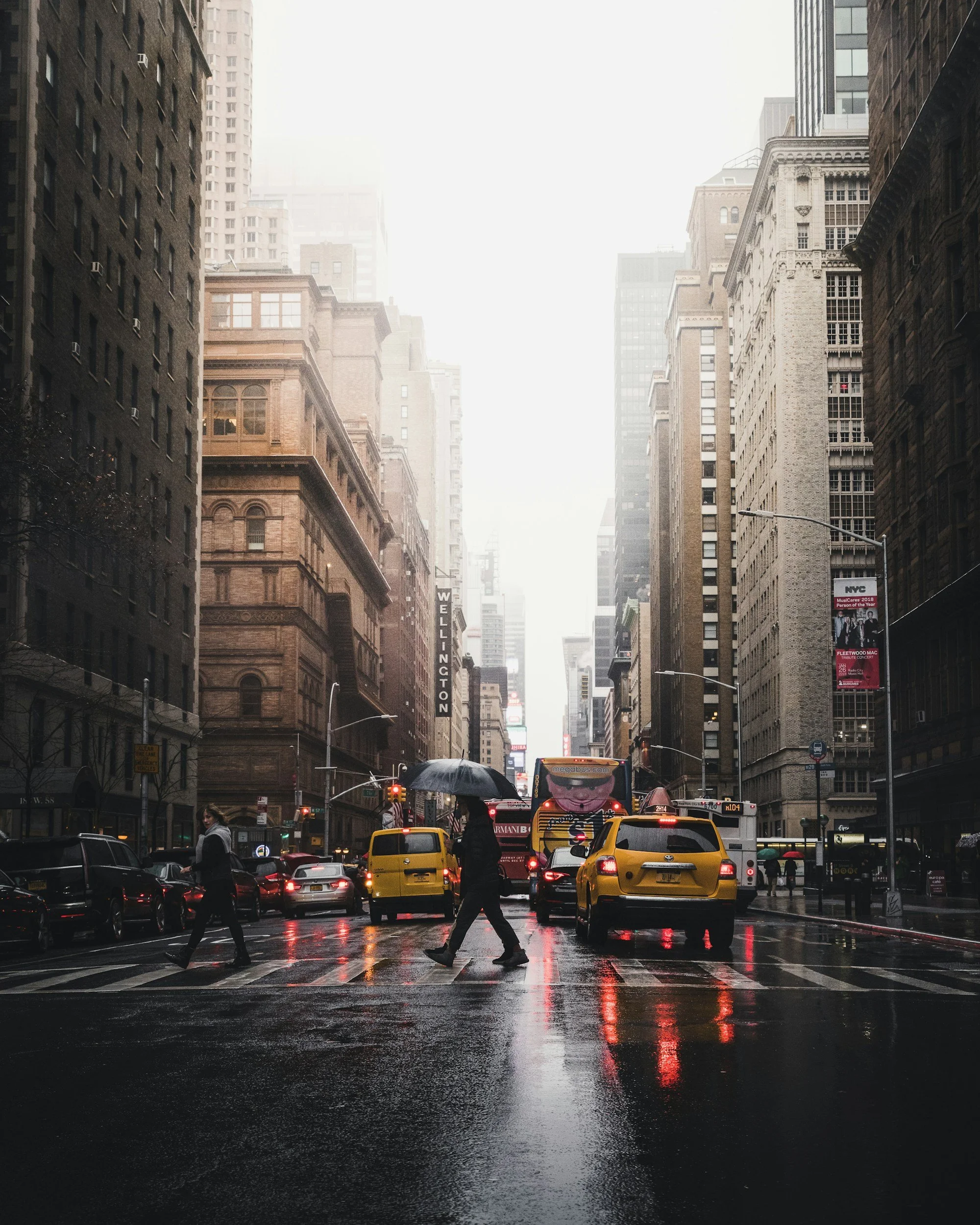 City street scene on a rainy day with tall buildings on both sides. Pedestrians cross a wet crosswalk, holding umbrellas, with traffic including yellow taxis and buses. The sky is overcast and the buildings are reflected in the wet pavement.