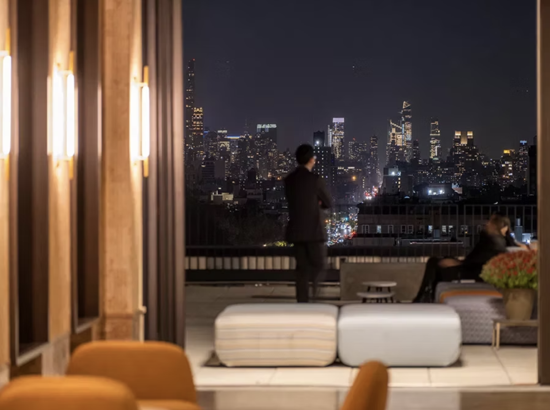 A view of a city skyline at night from a balcony. There is a man in a suit looking at the city, and a woman sitting and talking on the phone. The balcony is indoors with some furniture and curtains.