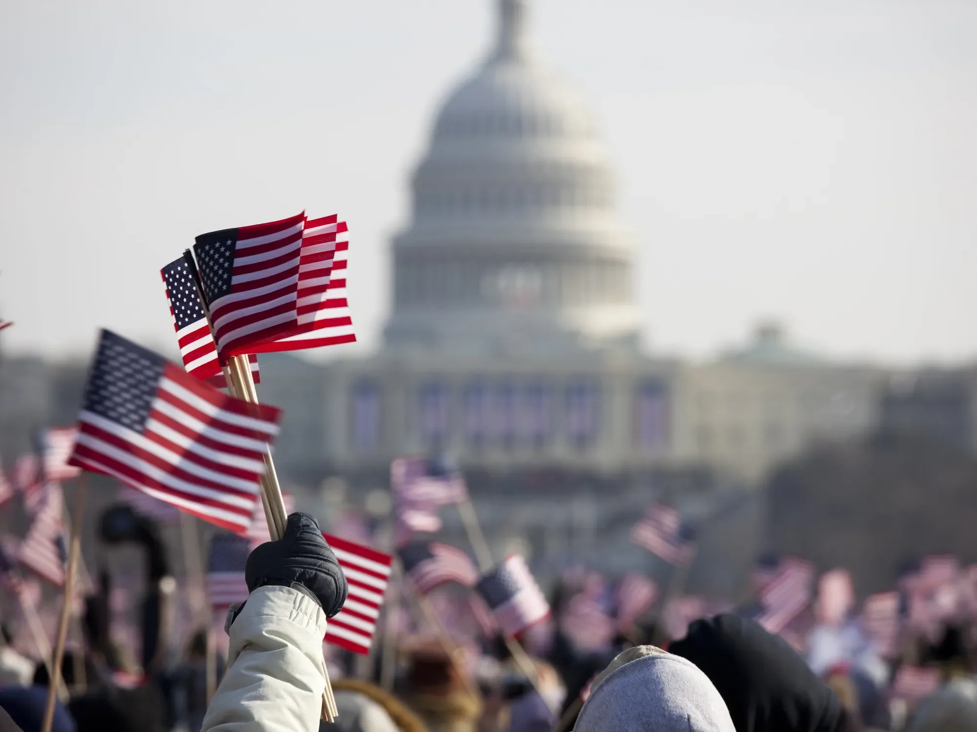 A person wearing a black glove and tan jacket holding small American flags amid a crowd, with the Capitol building in the background.