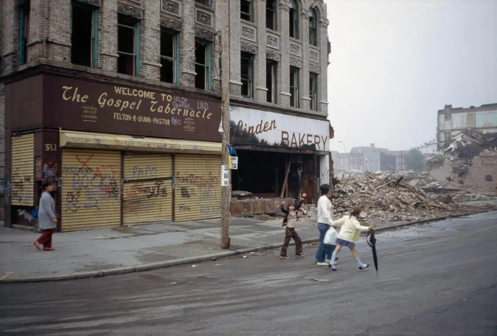 A city street with a damaged building and rubble debris on the side, three children and one adult walking across the street, with a bakery storefront and a boarded-up business with a graffiti-covered metal shutter.