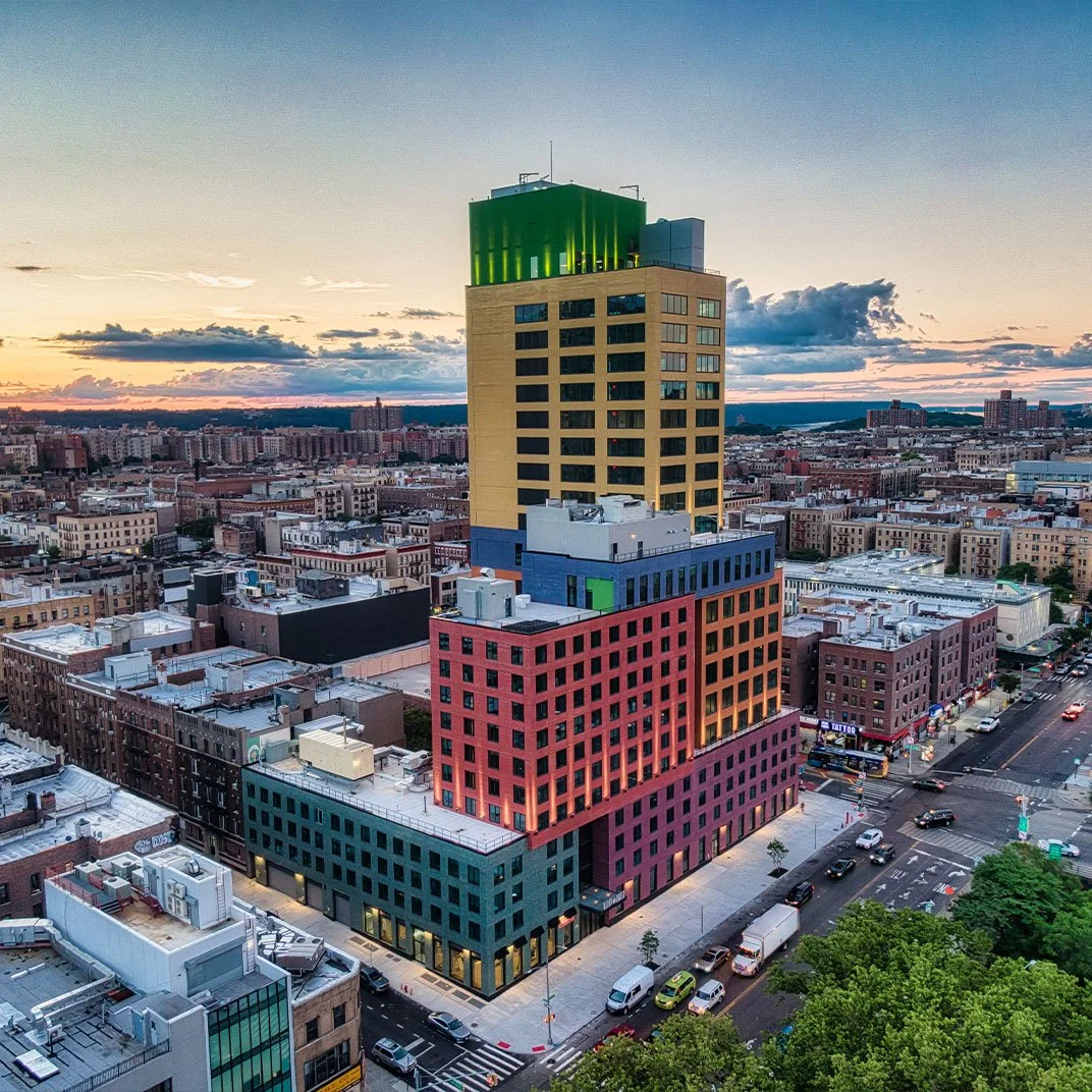 Colorful modern high-rise building with a yellow tower topped with a green structure, in an urban cityscape at sunset with numerous smaller buildings and streets.