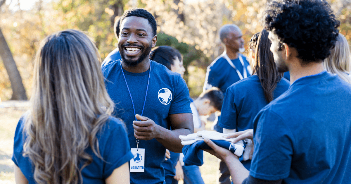 Group of volunteers wearing blue shirts outdoors, smiling and talking, with trees in the background.