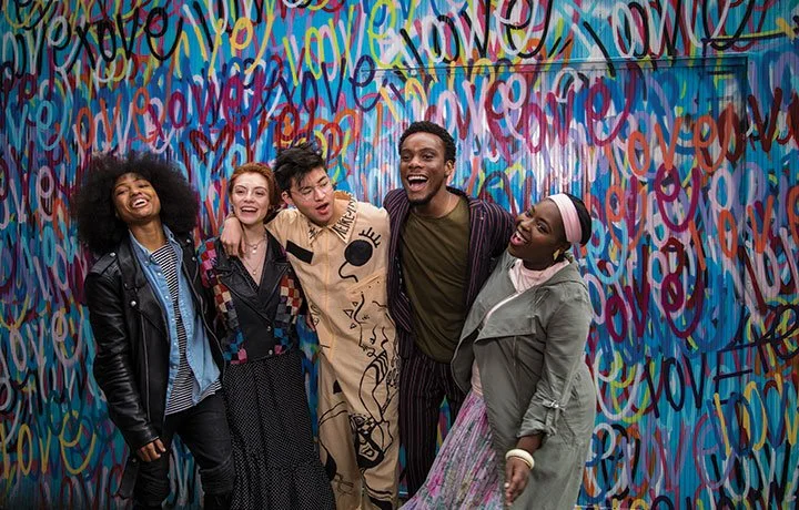 Five diverse young people smiling and laughing together, standing in front of a vibrant, colorful graffiti wall with the word 'love' repeatedly written in various styles.