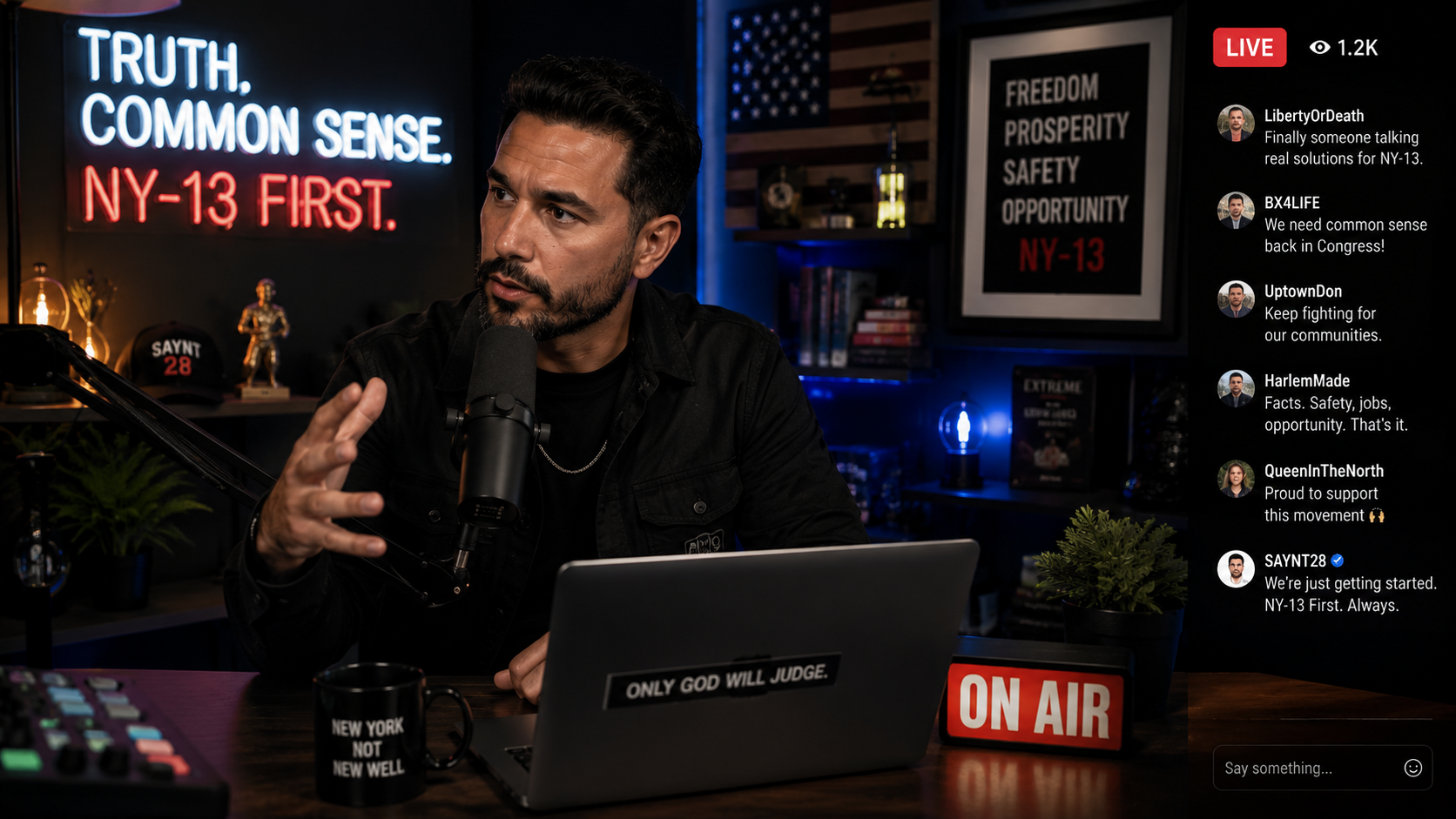 A man with a beard and dark hair speaks into a microphone during a live stream in a studio decorated with political campaign posters that say "TRUTH, COMMON SENSE. NY-13 FIRST" and "FREEDOM, PROSPERITY, SAFETY, OPPORTUNITY NY-13". The studio has American flags, books, and plants, with an "ON AIR" sign and a coffee mug on the desk.
