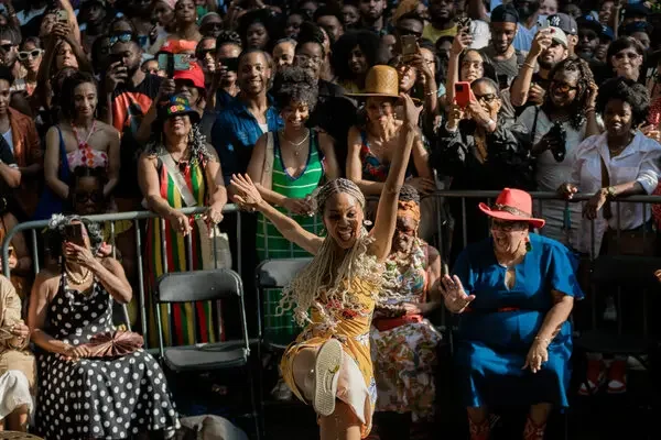 A woman dancing joyfully in front of an audience at an outdoor event, with onlookers smiling and capturing the moment on their phones.