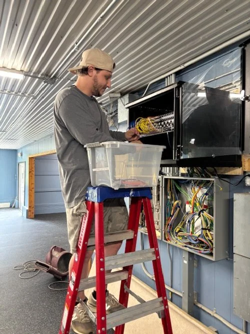 A man with a beard wearing a beige cap, gray shirt, and khaki shorts is standing on a red ladder, working on an open electronic control panel with colorful wires inside. The room has a metal ceiling and a large window or door with a black frame.