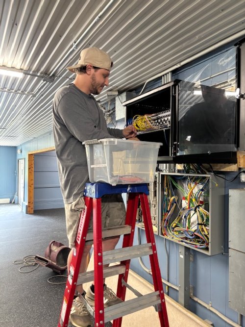 A man wearing a beige cap, a long-sleeve gray shirt, and beige shorts is standing on a red ladder, working on an electrical panel. There is a plastic bin with tools on top of the ladder. The workspace has a metal ceiling with visible support beams, a blue wall, and a large black screen or monitor on the wall.