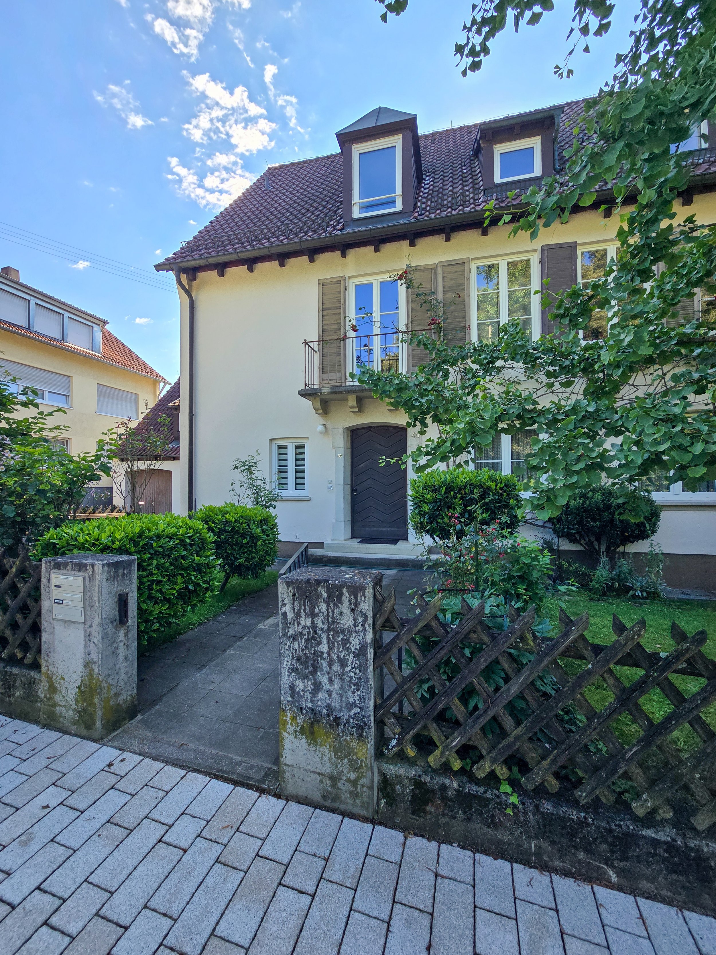 Ein zweistöckiges Haus mit weißen Wänden, dunklen Fensterläden und rotem Ziegeldach, umgeben von Grünanlagen und Zaun aus Holz und Beton