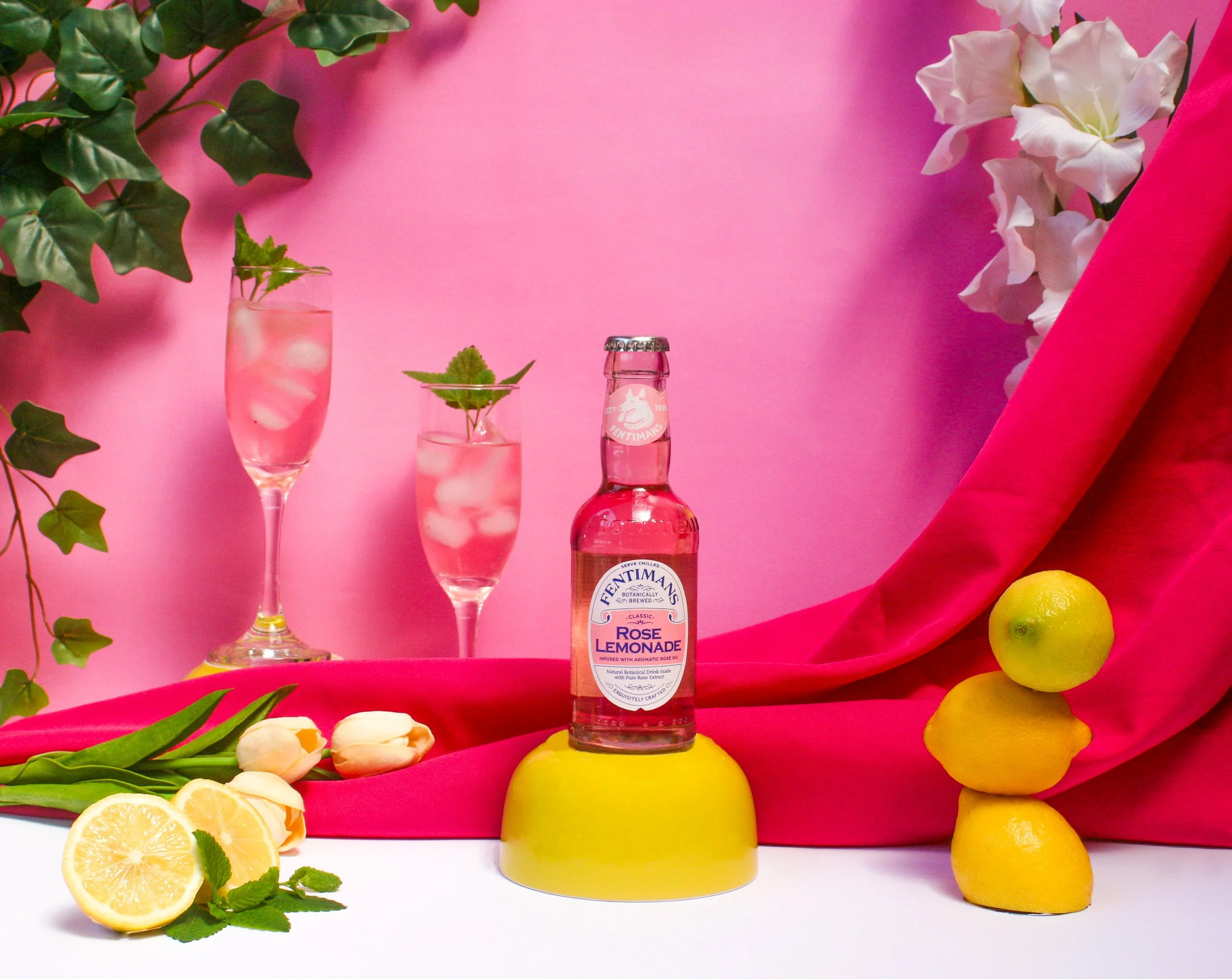 A still life of two glasses with pink beverages and ice, garnished with mint, a bottle of Fentimans Rose Lemonade between the glasses, and surrounded by lemons, tulips, and green leaves, with a pink backdrop and fabric draping.