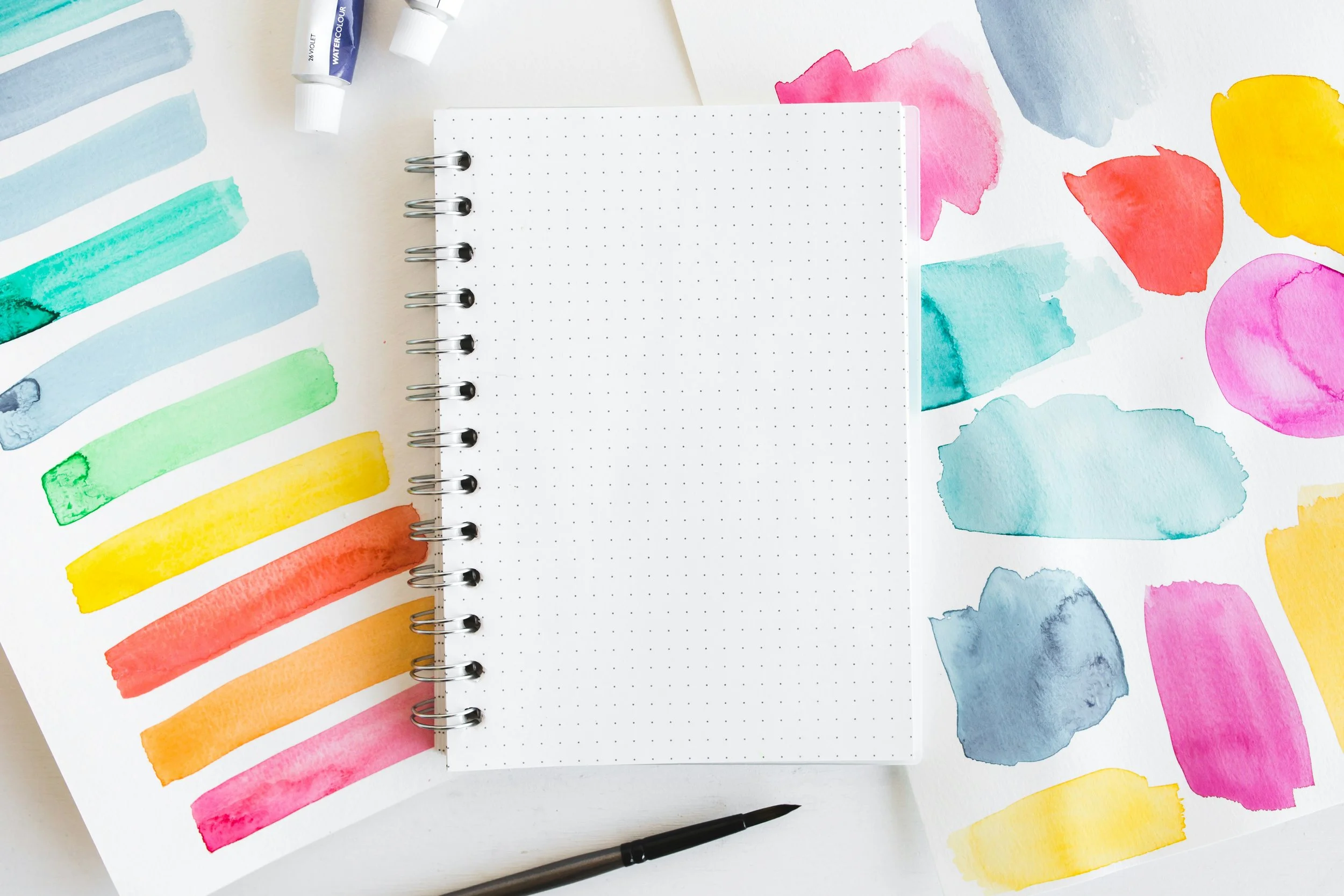 Watercolor paint swatches and a blank dotted notebook on a white surface.