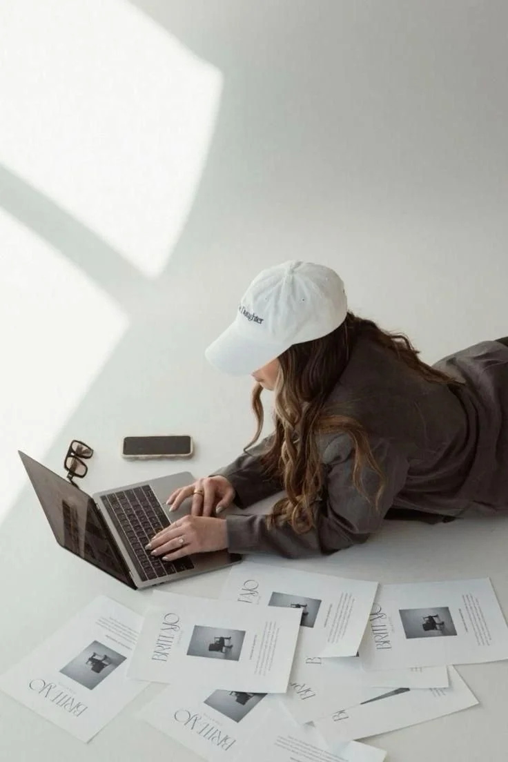 Woman lying on her stomach working on a laptop surrounded by printed documents, with a smartphone and glasses on a white table.