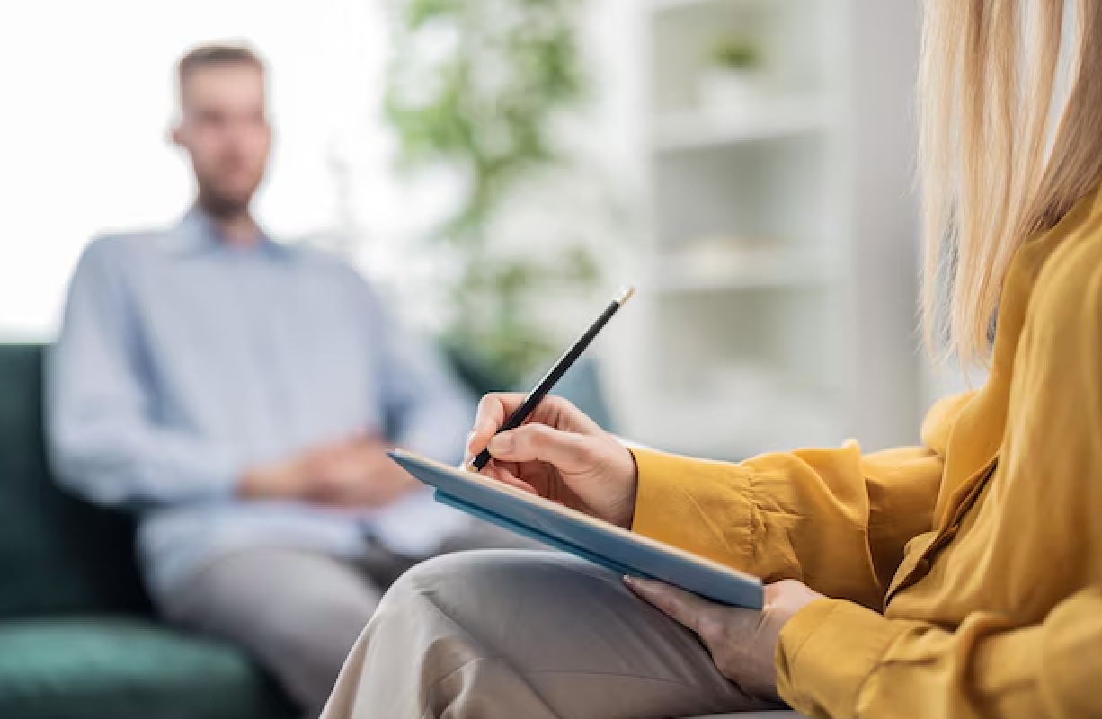 A woman in a yellow top is taking notes on a notepad with a pen while sitting across from a man in a light blue shirt during an interview in a bright office.