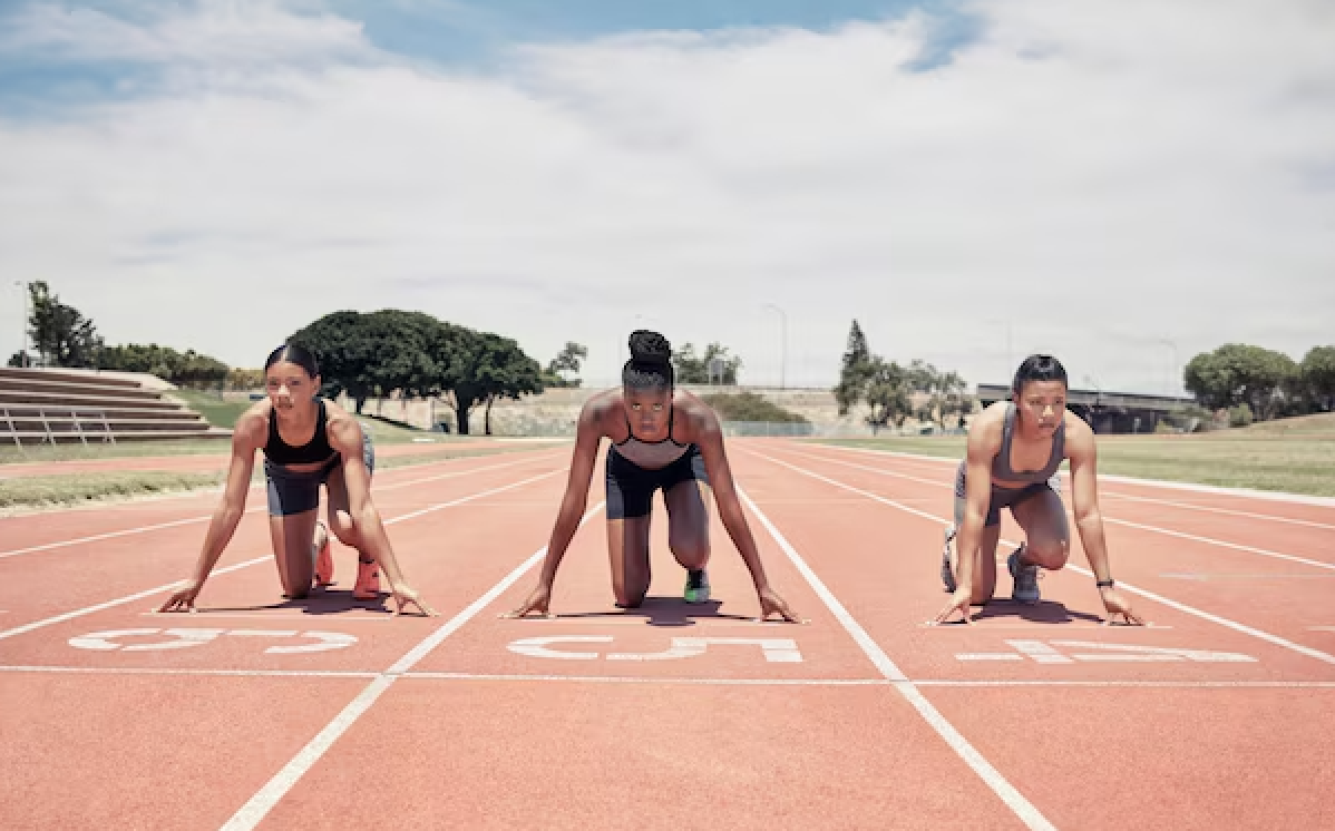 Three female athletes in starting positions on a running track, preparing for a race, outdoors with trees and a partly cloudy sky in the background.