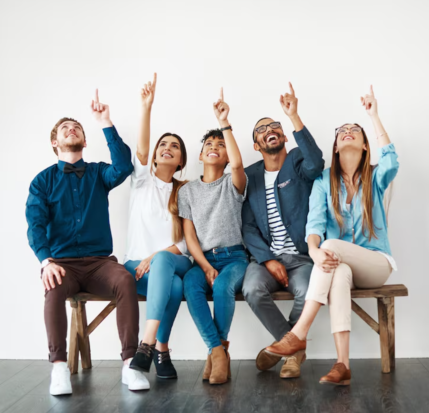 A diverse group of five young adults sitting on a wooden bench, looking up and pointing upward, smiling and appearing happy.