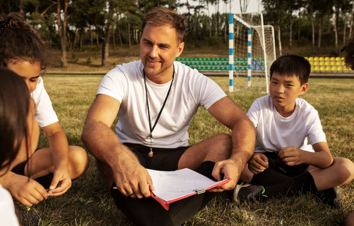 A man and children sitting on grass in a park, having a discussion, with soccer goals and trees in the background during daytime.