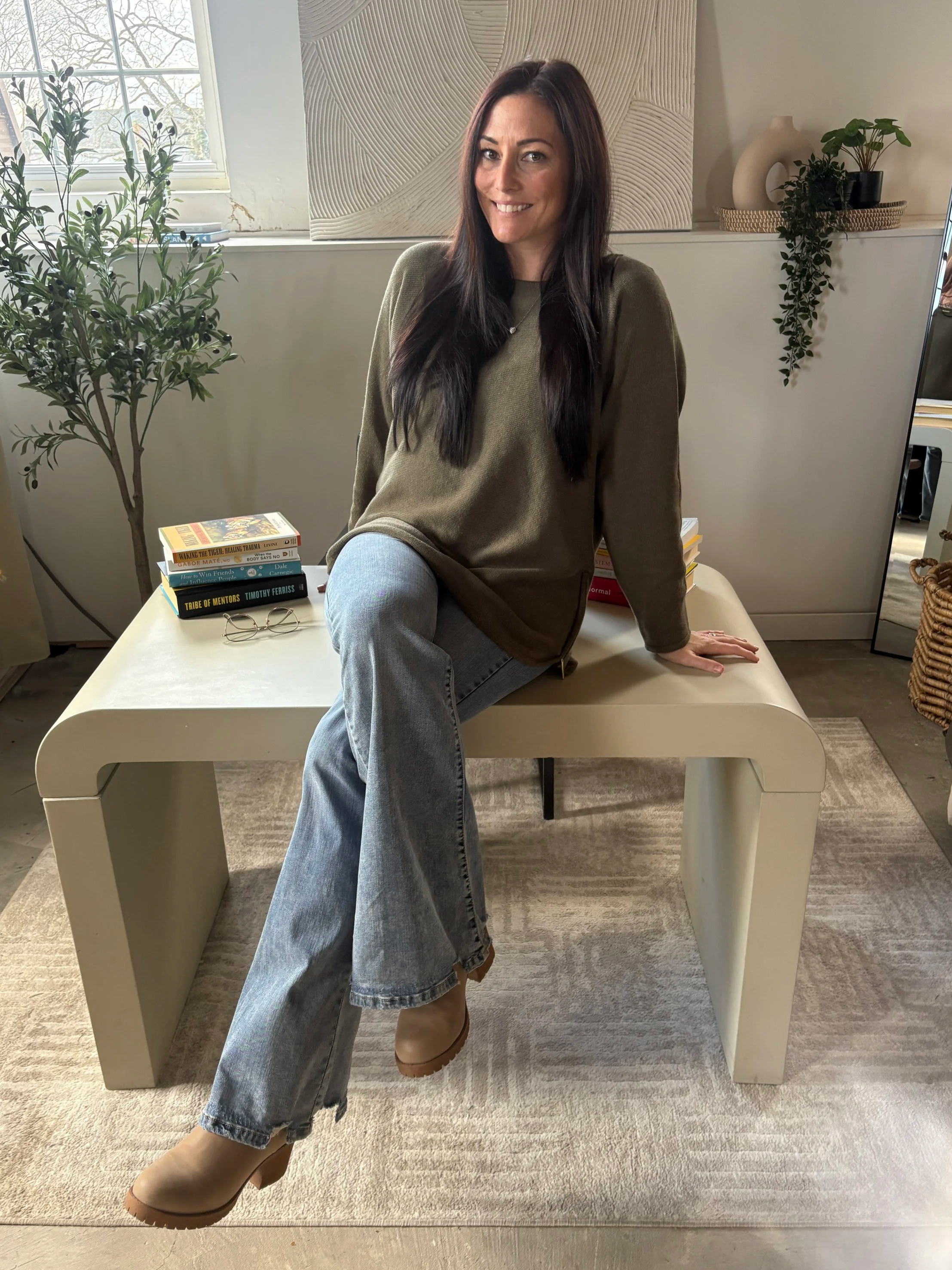 A woman with long dark hair and a smile sits on a modern beige bench in a bright room, wearing a green sweater, gray jeans, and beige boots. There are books, round glasses, a potted plant, and decorative objects on the bench and shelves behind her.