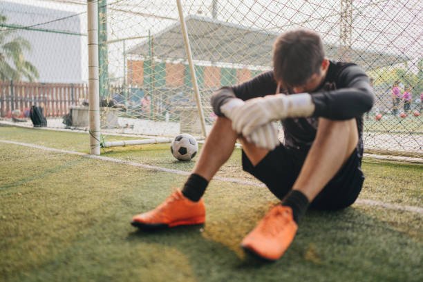 A young soccer goalkeeper sitting on the ground inside the goal, holding his leg in pain with a soccer ball nearby.