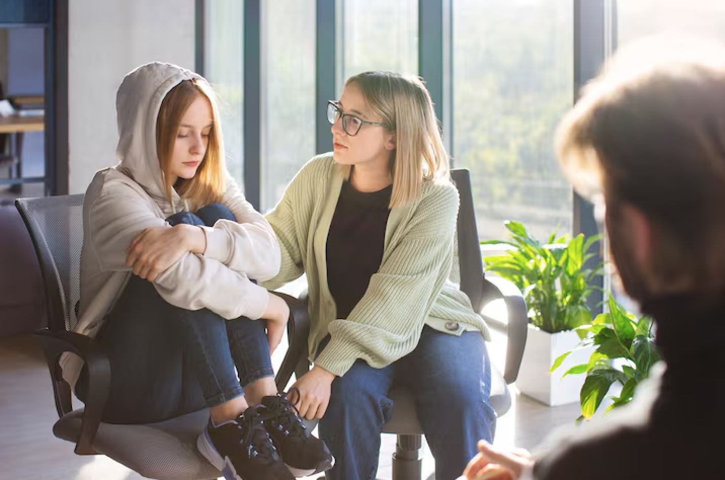 A young girl with a hoodie sitting on a chair, being comforted by a woman who appears to be her counselor or therapist, in a bright room with large windows and green plants.