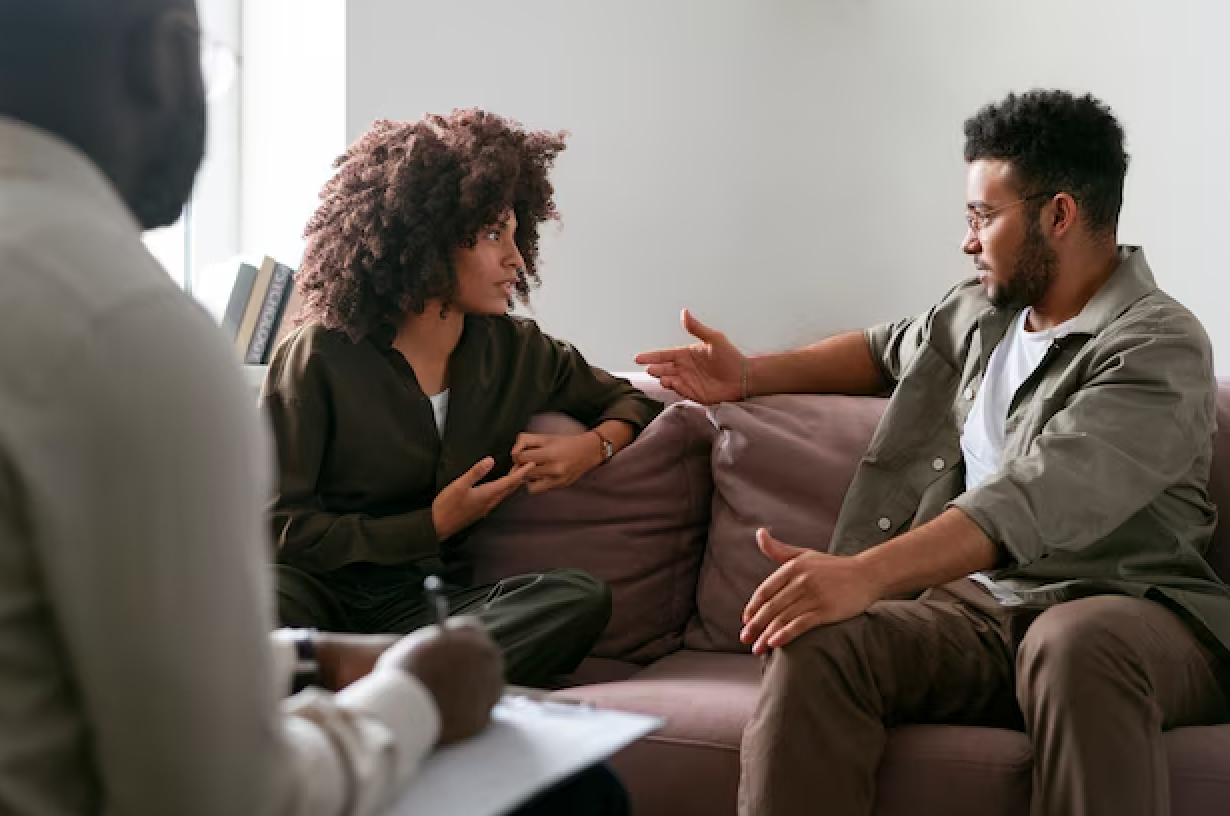 A woman and a man sit on a couch during a conversation, with a third person taking notes nearby, in a therapy or counseling session.