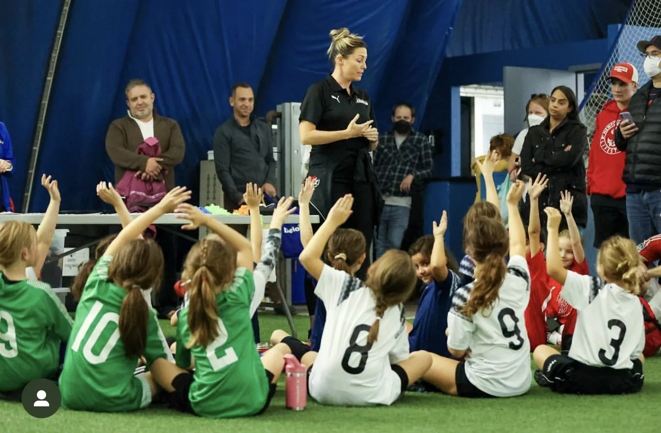 Young girls in sports jerseys sitting on indoor turf with their hands raised, listening to a woman speaking at a sports event, with several observers standing behind them.