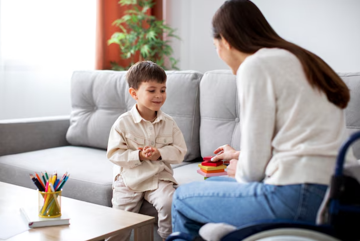 A woman in a wheelchair giving a stack of colored blocks to a young boy sitting on a gray couch in a bright room with a plant and curtain in the background.