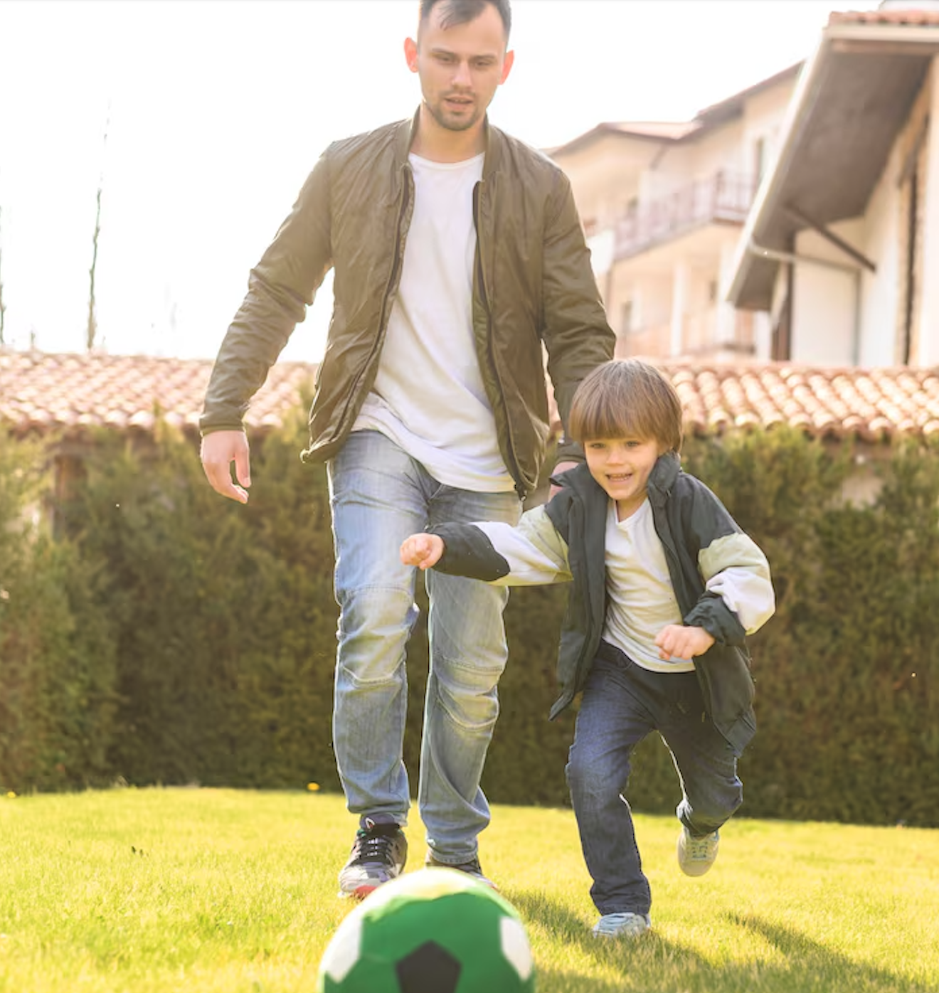A man and young boy playing soccer on a grassy yard, with the boy running towards a green and white soccer ball in front of him.