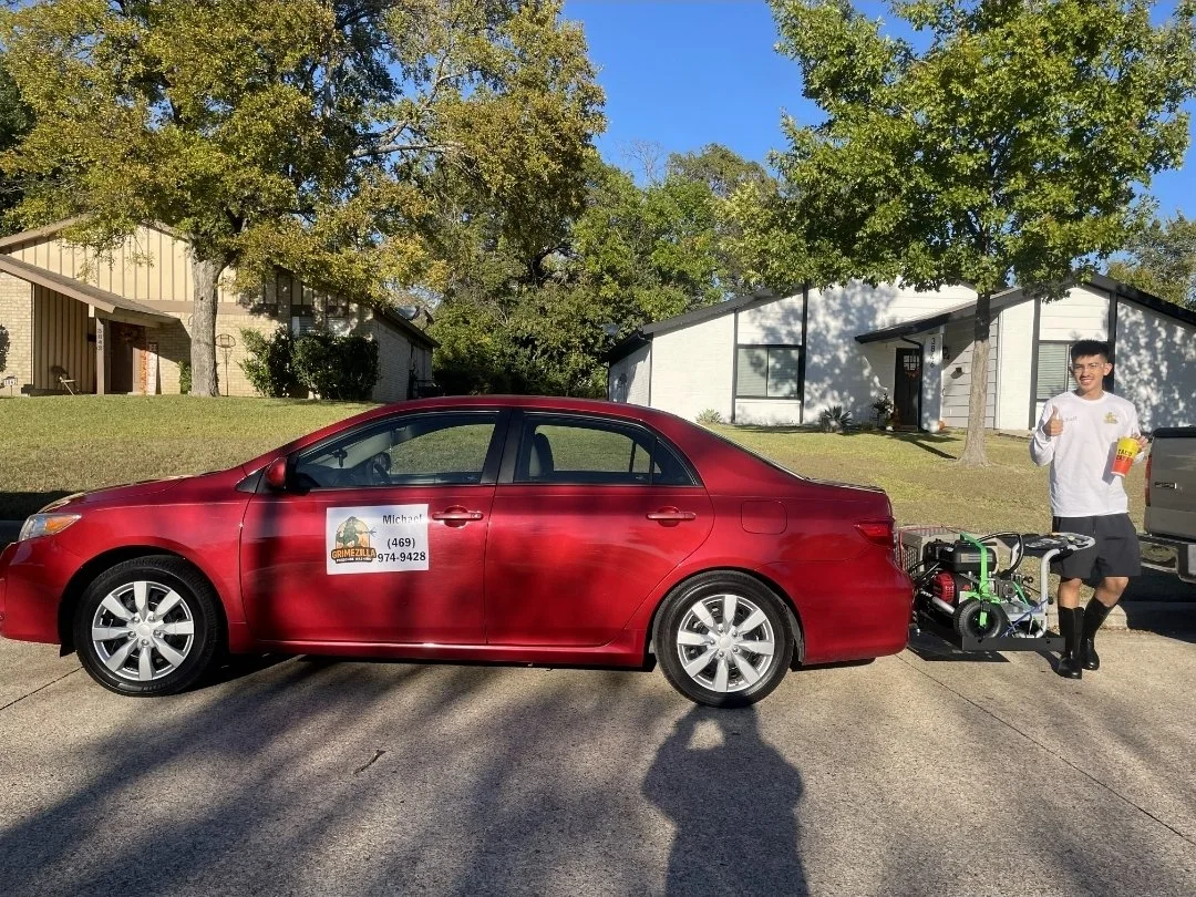 A red sedan parked on a residential street with a man standing next to it holding a snack and a thumbs-up sign. The car has a sign on the door advertising Michael's services with a contact number. The car has power washer attached to its rear.