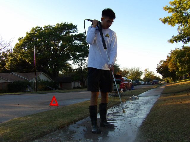 A young man in boots and shorts cleaning a sidewalk with a pressure washer in a residential neighborhood during the daytime.