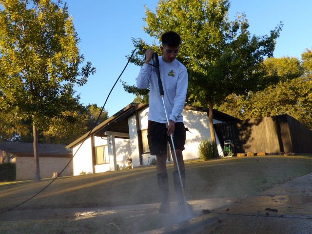 A young man pressure washing concrete with trees and houses in the background during daytime.