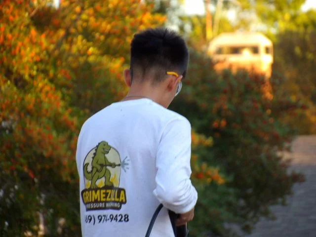 Back of a person wearing a white shirt with a cartoon Godzilla and the word 'Grimezilla' on it, outdoors in front of trees with autumn foliage.