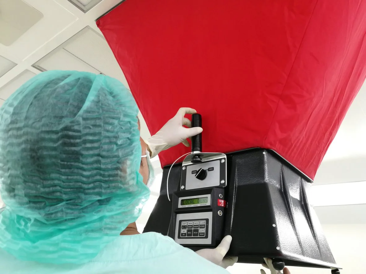 A healthcare professional in scrubs, a face mask, and a hair cover using a device with a digital screen and control panel to scan a red object in a medical or laboratory setting.