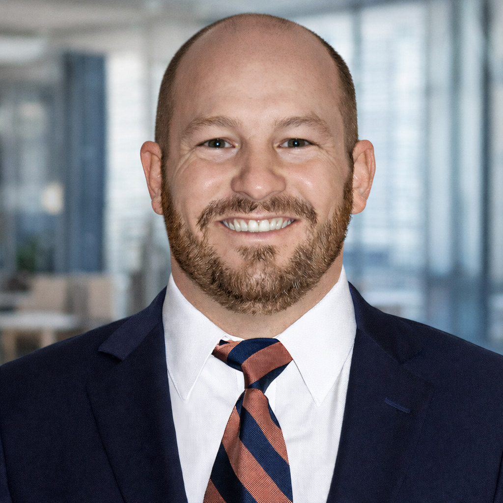 A man with a bald head and beard smiling in a professional suit with a striped tie, standing in a modern office environment.