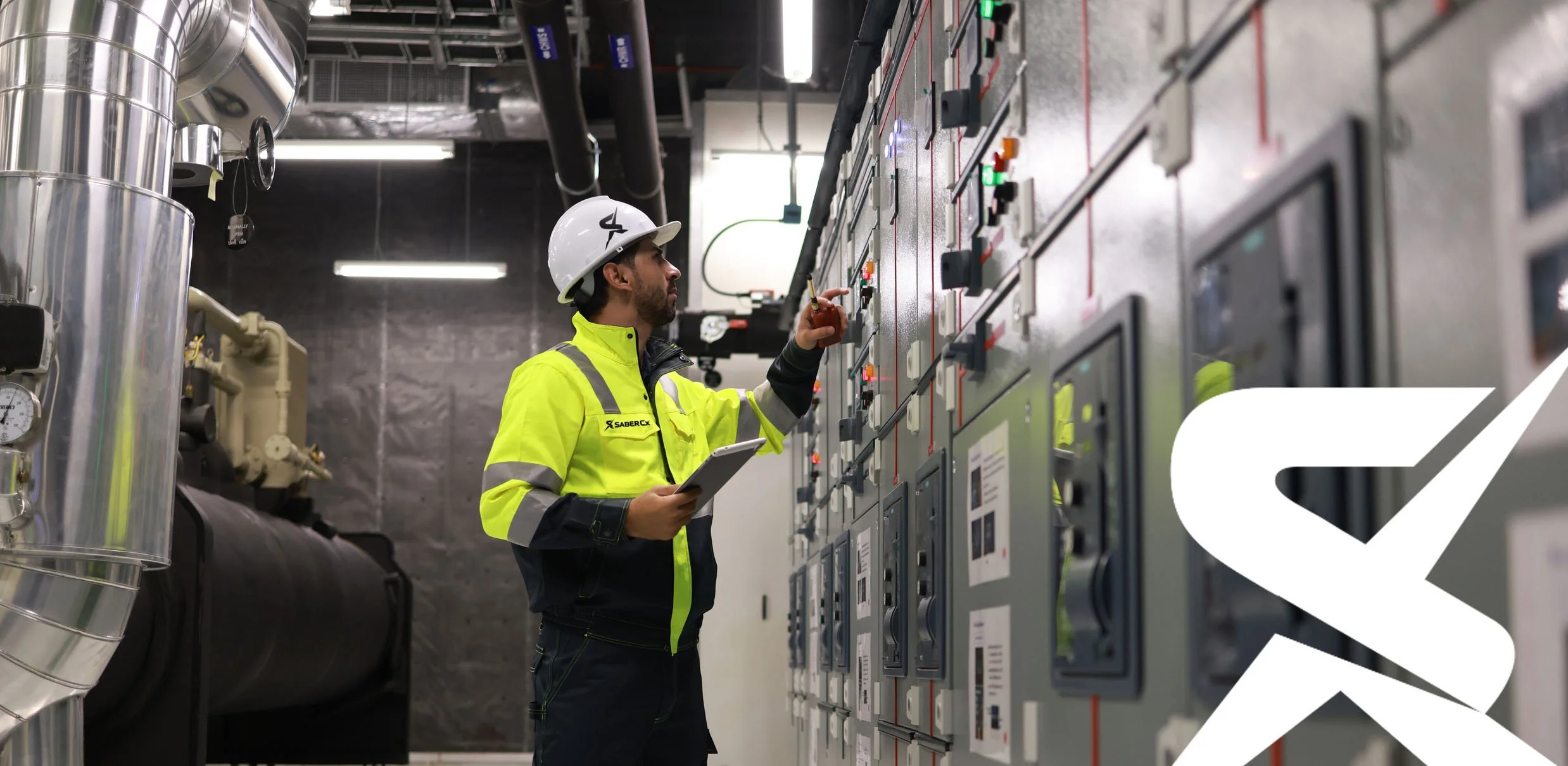 A male technician wearing a yellow safety jacket, a white hard hat, and black gloves, standing in front of an electrical control panel in an industrial facility, holding a tablet and adjusting controls.
