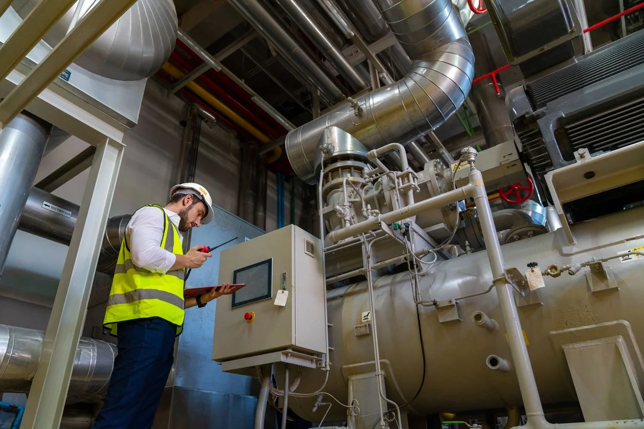 A man in a safety vest and helmet using a control panel in an industrial facility with large pipes and machinery.