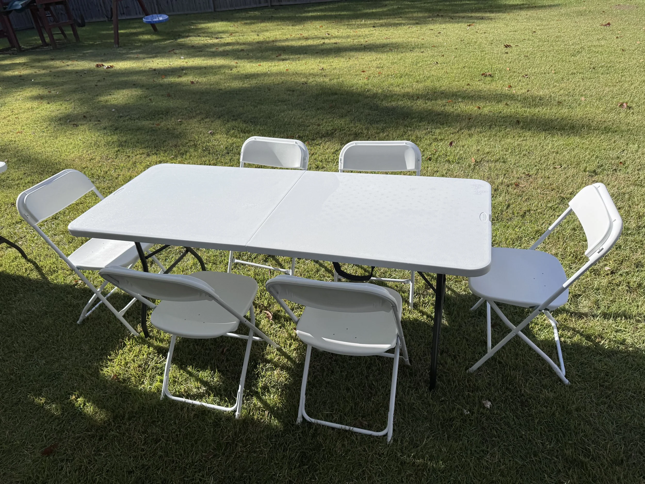 White plastic patio table and six matching white folding chairs outdoors on a grassy lawn, with some shadows and a fence in the background.